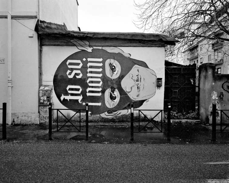 foto in bianco e nero con facciata di una casa con murales raffigurante uomo mascherato e scritta: io so i nomi