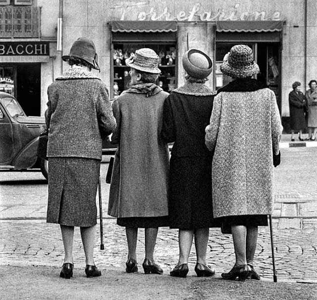 quattro donne sottobraccio di spalle con bastoni e cappelli che attraversano la strada