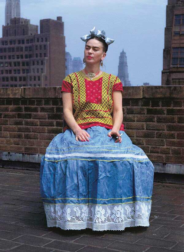 ritratto di Frida Kahlo seduta in terrazzo con gonna celeste maglia rossa e gialla fiocchi in testa e sigaretta in mano