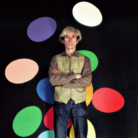 ritratto di Andy Warhol con le braccia incrociate davanti a sfondo nero con macchie ovali colorate