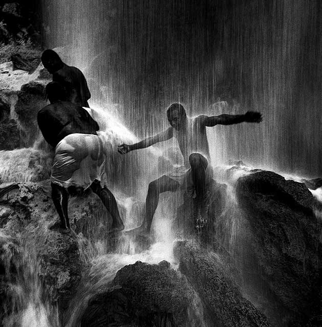 fotografia in bianco e nero di tre ragazzi seduti sugli scogli sotto l'acqua di una cascata