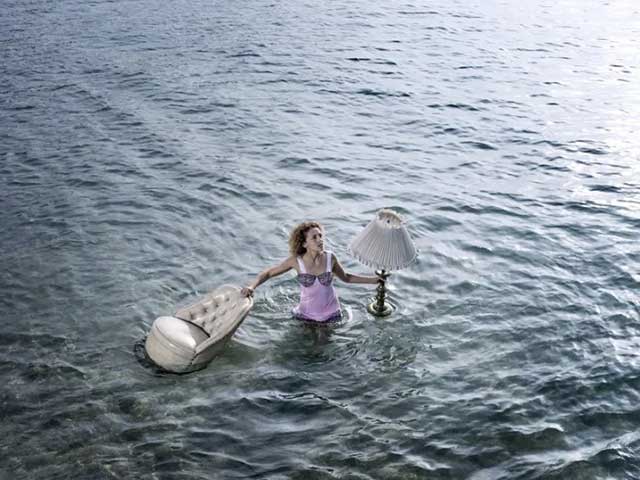 fotografia a colori di una Donna in mare vestita di rosa con in mano un lampadario e una poltrona