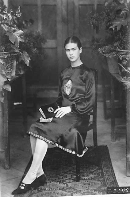 ritratto in bianco e nero di frida kahlo giovane con vestito nero,capelli raccolti,calze bianche e libro in mano seduta su una sedia
