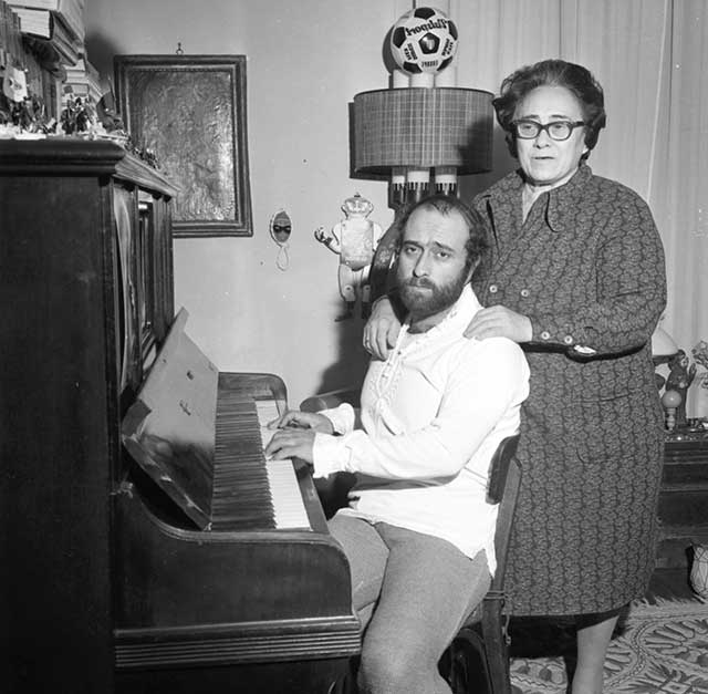 fotografia in bianco e nero di Lucio Dalla al pianoforte con dietro la mamma che gli tiene le mani sulle spalle