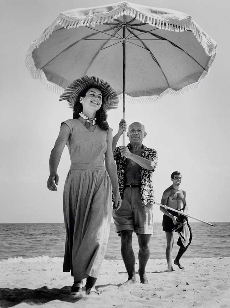 Pablo Picasso cammina sulla spiaggia tenendo l'ombrello alla moglie