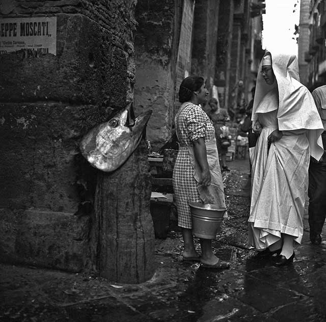 donna con vestito a quadri e grembiule con secchio in mano che parla con una suora per strada secchio ch