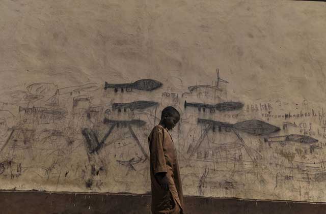 bambino africano che cammina per strada con dietro muro con disegni di mitra