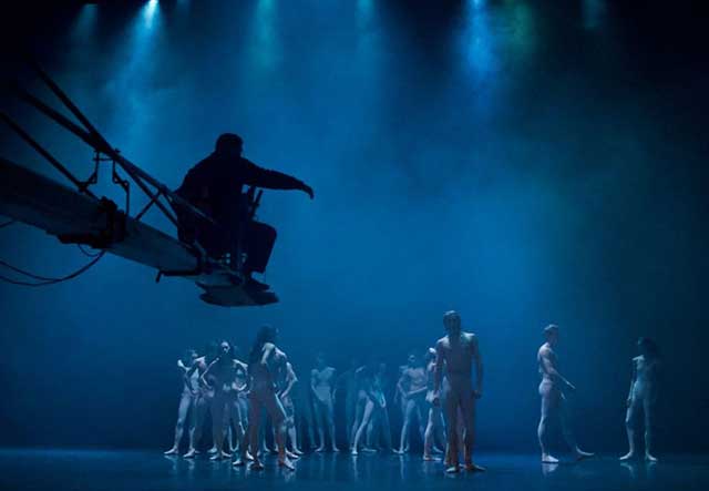 immagine con luci blu di ballerini sul palco con sagoma nera di operatore con di macchina da presa che da indicazioni dall'alto