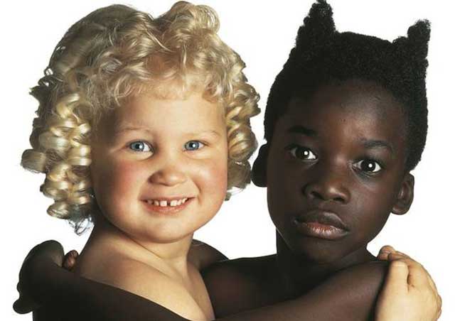 primo piano di bambina bianca con riccioli biondi e occhi azzurri abbracciata a bambina nera con capelli e occhi scuri