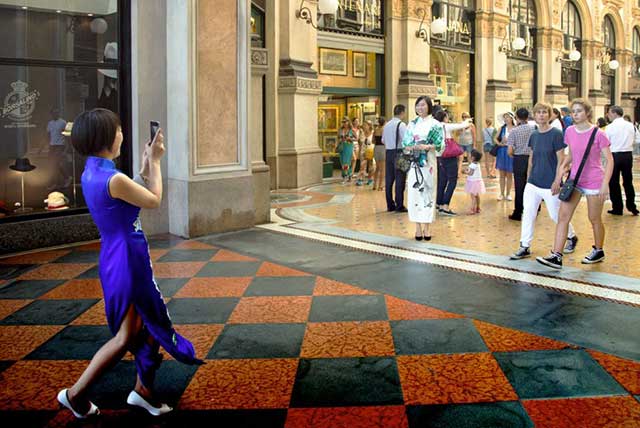 ragazza giapponese con vestito blu lungo con spacco laterale e scarpe bianche che scatta foto con il telefonino ad altra ragazzasorridente con vestito bianco e scarpe nere dentro galleria affollata