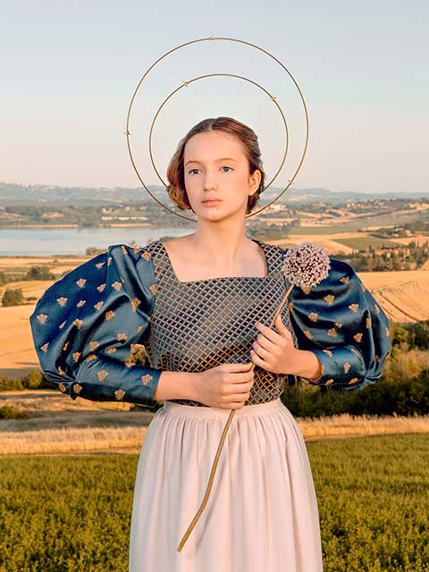 ragazza con aureola capelli raccolti vestito blu con grandi maniche a palloncino e gonna bianca che tiene fiore in mano davanti a paesaggio di campi con lago sul fondo