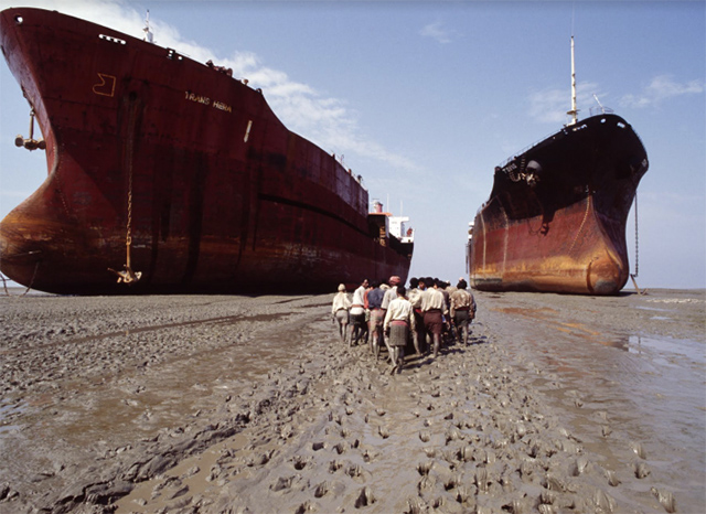uomini camminano scalzi sulla sabbia in mezzo a due enormi navi arrugginite