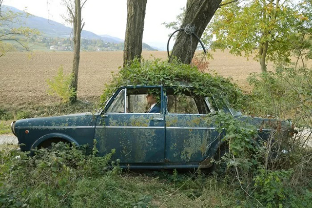 Ricarica Foto Festival 2022 uomo in un bosco dentro una macchina vecchia con la vernice scolorita e ricoperta di edera
