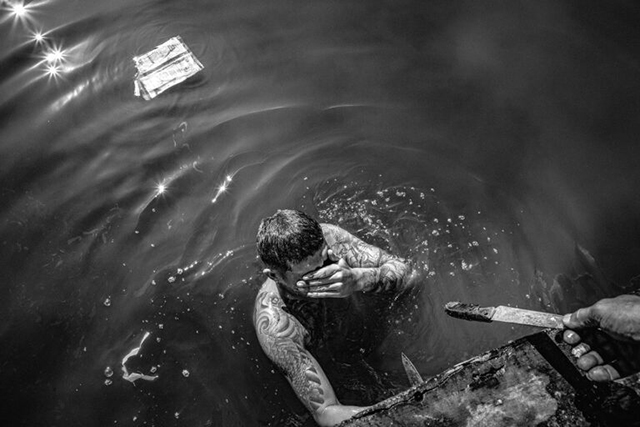 TERRA! FESTIVAL DI FOTOGRAFIA E CINEMA uomo in acqua con le braccia ricoperte di tatuaggi che con una mano si asciuga il viso e con l'altra si appoggia ad un muretto dove spunta una mano che gli porge un coltello