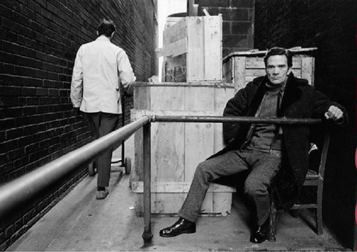Pier Paolo Pasolini Sotto gli occhi del mondo foto in bianco e nero di Pier Paolo Pasolini seduto su una sedia in strada vestito elegante con completo scuro e cappotto nero