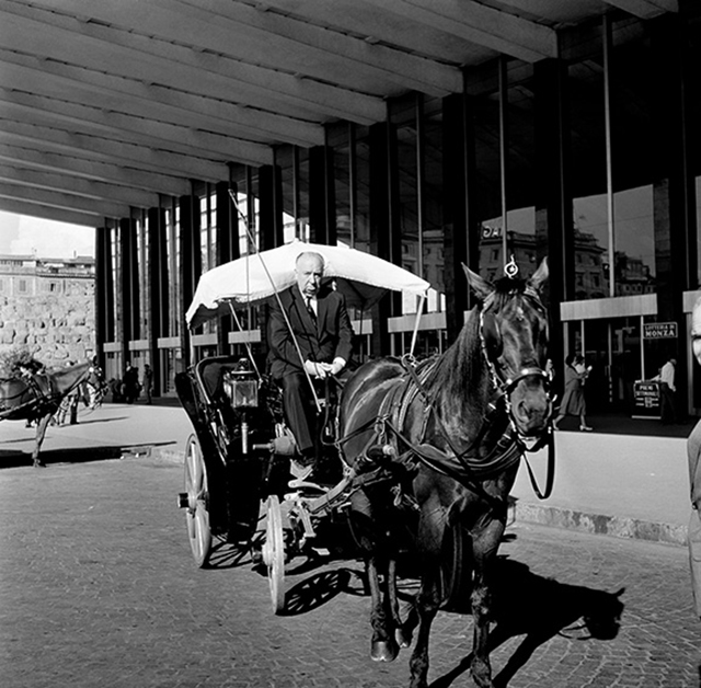 La memoria delle stazioni foto in bianco e nero con carrozza trainata da cavallo nero con sopra il regista Alfred Hitchcock al posto del conducente che tiene le redini in completo nero con giacca e cravatta davanti all'entrata della stazione Termini