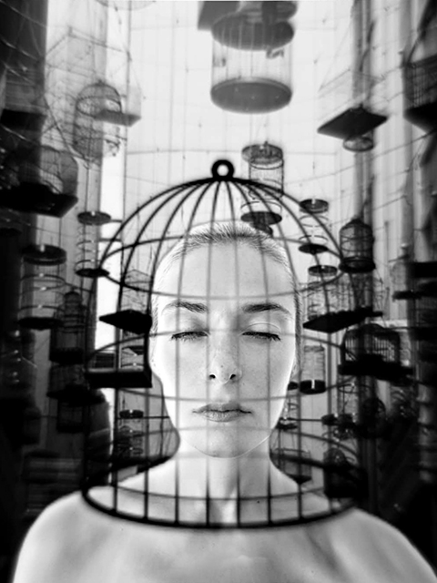 Lidia Musso primo piano di donna con gli occhi chiusi i capelli raccolti e la testa dentro una gabbia di uccellini a torso nudo con sullo sfondo altre gabbie appese