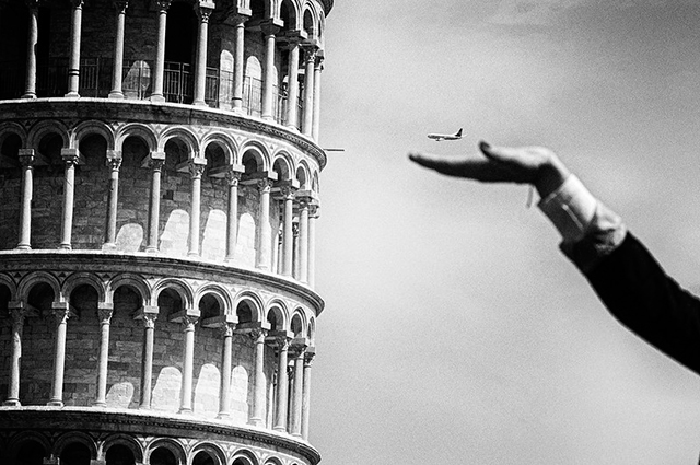 Pisa Street Photography dettaglio della torre di pisa con in primo piano braccio con la mano tesa come a prendere aereo che passa nel cielo