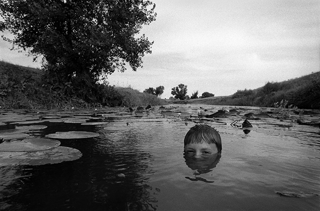 Festival Fotografia Etica foto in bianco e nero con in primo piano bambino biondo sorridente con il viso mezzo coperto dall'acqua mentre fa il bagno in un laghetto fra alberi e prati in lontananza