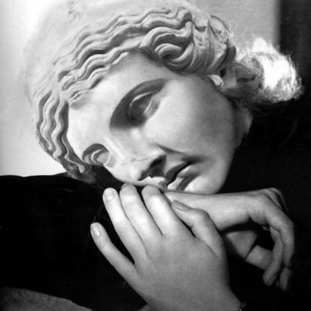Photo Open Up 2022 foto in bianco e nero con primo piano di donna con testa reclinata sopra il braccio,le mani incrociate davanti al viso coperto da una maschera da statua greca