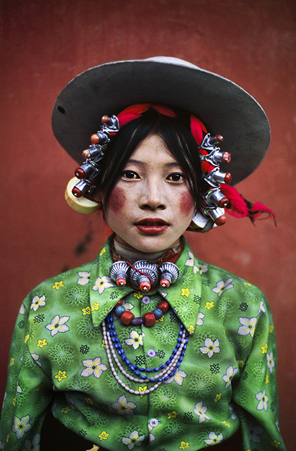 Steve McCurry Texture primo piano di ragazza peruviana con cappello bianco tondo da cui escono dei pendagli argentati arrotolati in un foulard rosso camicia verde con fiori gialli e bianchi allacciata fino al collo e sei collane di varie misure al collo