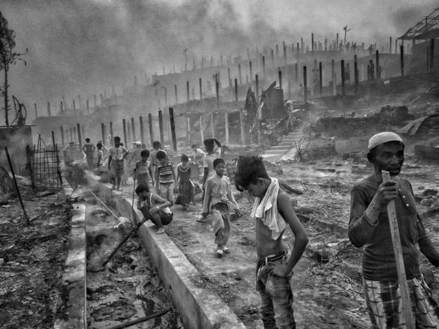 Un ambiente ostile: vita dei Rohingya immagine in bianco e nero di campo profughi con bambini uomini e donne che camminano in mezzo a macerie fango e fumo