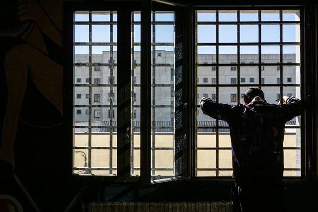 ri-scatti .per me si va tra la perduta gente. uomo in controluce nella cella di un carcere appoggiato con le braccia alzate ad una finestra con le grate all'esterno un cortile ed un altro edificio