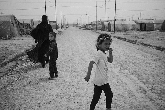 christian tasso e alessio romenzi the thine line foto in bianco e nero con due bambini e una donna coperta da velo nero che camminano per una strada deserta polverosa con alcune tende in lontananza