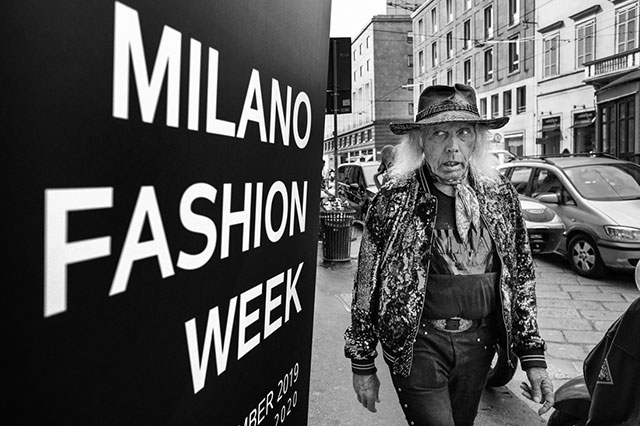 cities foto in bianco e nero con persona anziana con cappello da cow-boy e giubotto di palettes cammina per strada guardando con espressione strana il cartello della milano fashion week