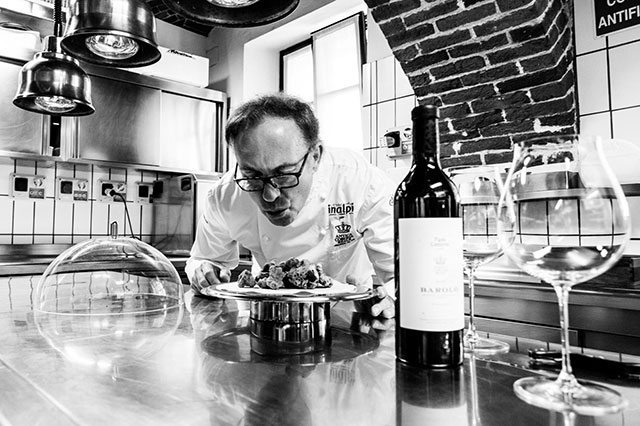 Fabio Bonanno foto in bianco e nero di chef nella sua cucina che soffia su piatto di pietanza appoggiato sul bancone con davanti bottiglia di vino e bicchiere
