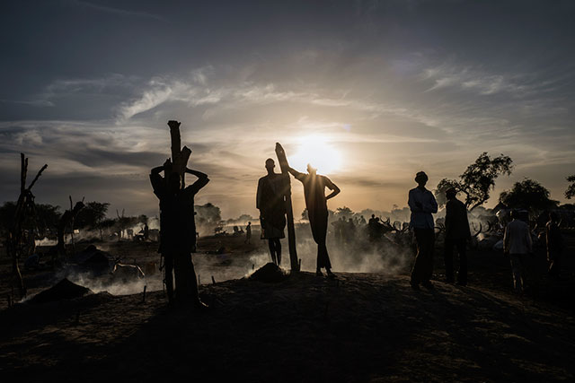 focus on future 3 uomini appoggiati a dei lunghi bastoni in controluce al tramonto con alle spalle altri uomini e donne e animali in mezzo al fumo