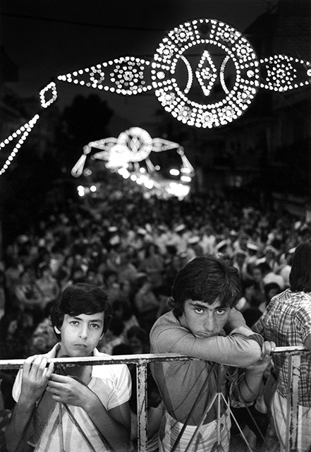 l'italia di Magnum da robert capa a paolo pellegrin due ragazzi appoggiatiad una ringhiera con facce tristi con alle spalle molta gente che partecipa a festa di paese con tante luci appese per la strada