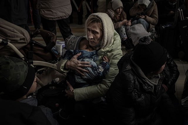 narnimmaginaria fotografia a colori di donna con l'espressione preoccupata seduta in terra che stringe un bimbo piccolo circondata da altre persone sedute