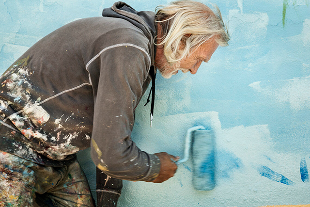 Ilaria Ranalletta I sogni li spendo per strada Uomo con i capelli lunghi bianchi e giacca marrone dipinge per strada muro con pittura celeste con rullo