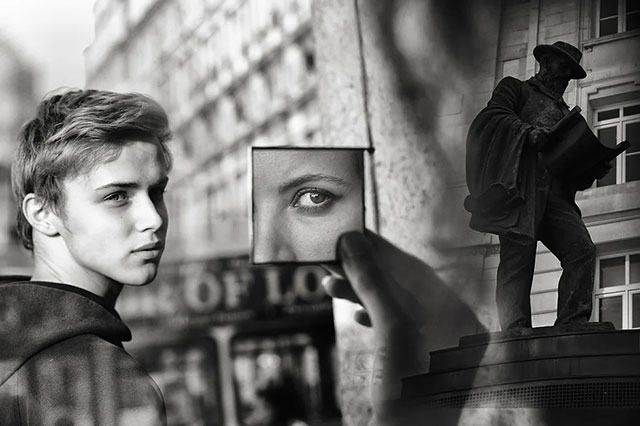 Med Photo Fest 2022 foto in bianco e nero di primo piano di ragazzo biondo dietro una vetrina dove c'e una mano che tiene uno specchietto con riflesso in occhio