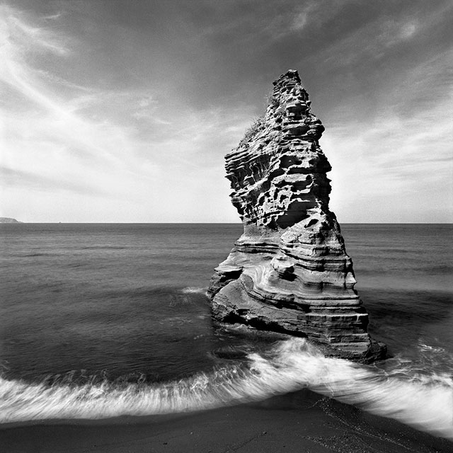 Mimmo Jodice Abitare metafisico immagine in bianco e nero di grande scoglio circondato dal mare con un onda bianca che sta per infrangersi sopra