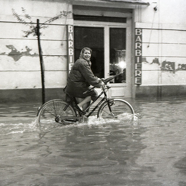 foto riccione immagine in bianco e nero di donna sorridente che va in bicicletta passando davanti a negozio di barbiere