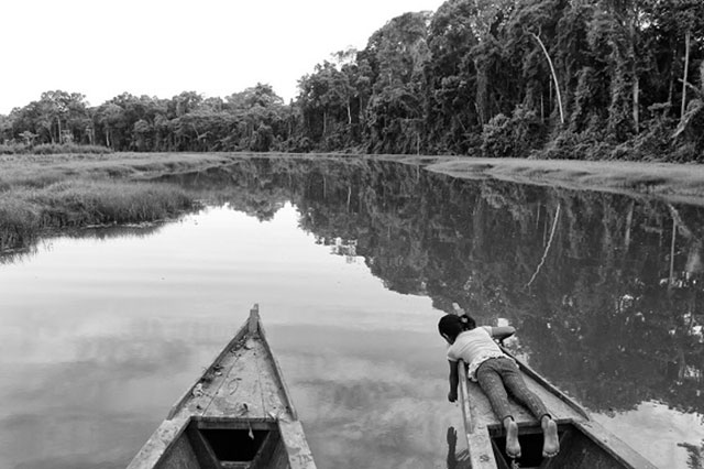 Pino Ninfa Entrada Proibida. Cronache Amazzoniche foto in bianco e nero con la punta di due barche che navigano su un fiume in Amazionia,sulla barca a destra una una bambina sdraiata che si sporge con un braccio a toccare l'acqua