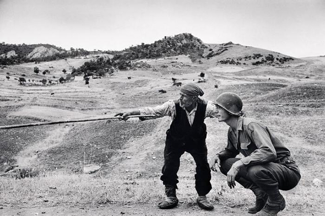Robert Capa L'opera 1932-1954 piccolo uomo anziano vestito con gilet nero camicia bianca fazzoletto in testa e pantaloni neri indica con un bastone la strada a soldato accovacciato accanto a lui