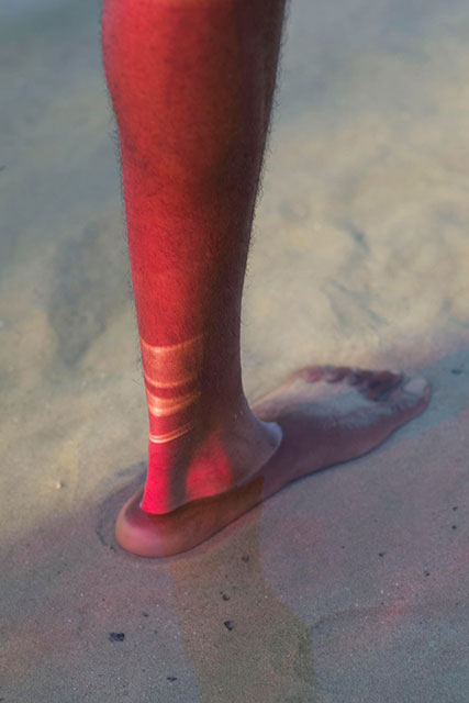 On the verge foto a colori di gamba di uomo illuminata da luce rossa con strisce piu chiare sul tallone e piede immerso in acqua marina trasparente e sabbia sul fondo