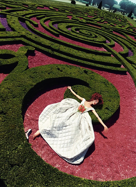 David LaChapelle Milano foto a colori di modella vestita di biancoj con fiori rossi sul petto sdraiata in giardino con terra rossa e siepi a forma di labirinto