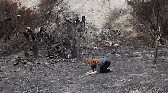 fatma bucak Torino foto a colori di donna inginocchiata su terra grigia intorno a rocce e alberi bruciati