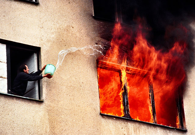 Livio Senigalliesi Milano foto a colori di uomo che dalla finestra con un secchio lancia acqua nella finestra accanto dalla quale escono le fiamme