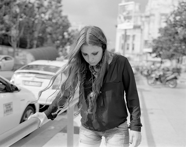 Mark Steinmetz foto in bianco e nero di ragazza giovane con capelli castani lunghi maglia nera e jeans che guarda in basso appogiata a scorrimano mentre cammina per la strada
