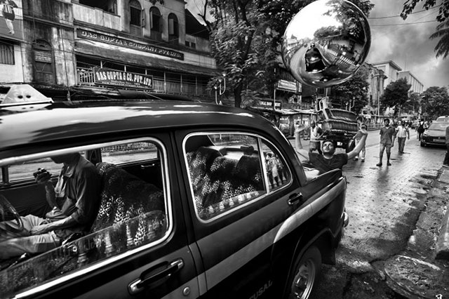 Max Vadukul Milano foto in bianco e nero con macchina parcheggiata in strada dalla quale si intravede una persona.dietro la macchina un ragazzo che fa volare un pallone specchiato