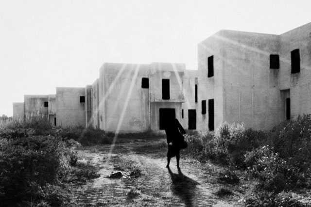 Nerina Toci foto in bianco e nero di figura di donna in controluce che cammina su vialetto erboso fra case bianche abbandonate con finestre nere e raggio di luce che filtra dal cielo sopra i tetti
