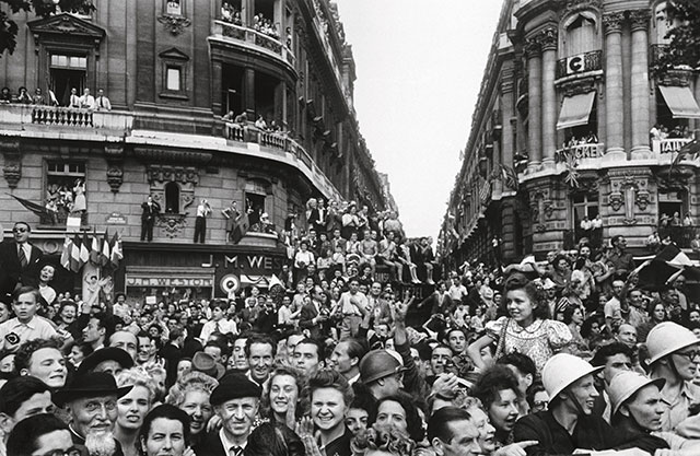 robert capa nella storia mostra mudec milano folla di gente sorridente per la strada davanti a due palazzi con gente affacciata e bandiere appese