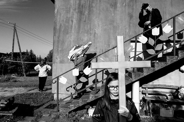 eyeshot roma foto in bianco e nero di bambina con croce di legno in mano dietro di lei un prete e un uomo con una statua di una madonna in mano che scendono le scale e una donna di spalle con le braccia sui fianchi