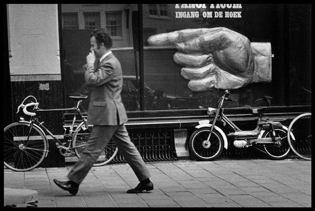 elliott erwitt abano terme foto in bianco e nero di uomo che cammina per la strada con un dito nel naso davanti a cartellone con grossa mano che indica verso di lui