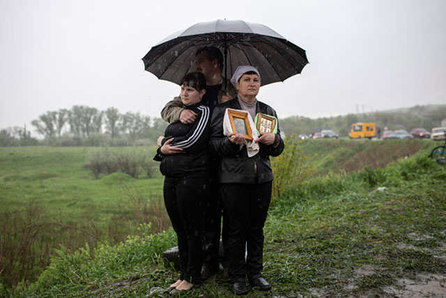 Francesca Volpi Brescia foto a colori di due donne e un uomo sotto un ombrello mentre piove tengono in mano delle fotografie incorniciate